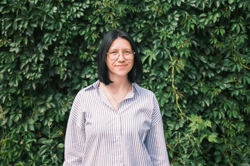 A beautiful young woman with black bob hair, wearing glasses and a light striped summer shirt, stands against the background of green grape leaves.