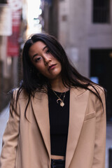 Stylish young asian woman confidently posing in a beige blazer and gold necklaces against a city backdrop