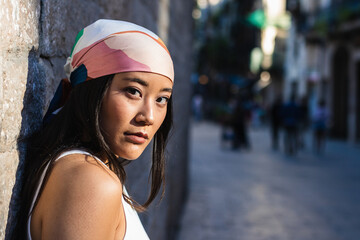 Confident young asian woman with headscarf leaning against stone wall in busy city street: a...