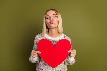 Charming young woman holding red heart symbol on khaki background, promoting love and affection with a playful kiss