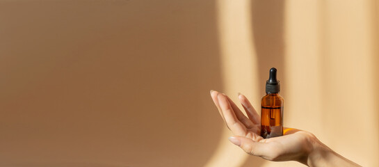 Bottle of serum in women's hands. Glass bottle with dropper cap on a beige banner with a copy space. Unmarked Amber container in sunlight