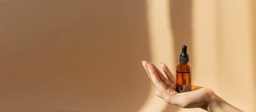 Bottle of serum in women's hands. Glass bottle with dropper cap on a beige banner with a copy space. Unmarked Amber container in sunlight