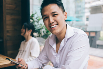 Obraz premium Asian smiling young man with smartphone sitting at desk with laptop