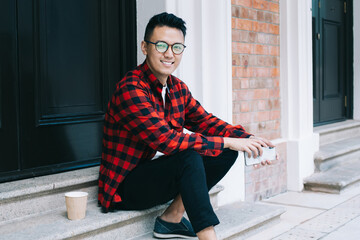 Young ethnic man in casual clothing smiling while sitting on doorstep