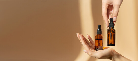 Women's hands hold bottles of amber glass with cosmetic serum on brown background. Mockup of container with dropper lid with collagen in rays of sunlight. Copy space,banner