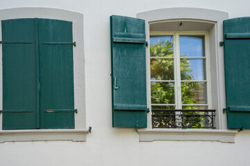 Two windows in the wall of a house with old fashioned green window shutters. One shutter is open, one closed. Reflection of trees and blue sky in the windows of the open shutter.