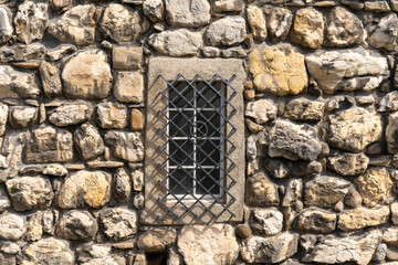 Texture of old stone wall with big boulders of limestone in different pastel colors. In the wall an old fashioned barred window with a metal grid with diagonal metal bars is inserted.