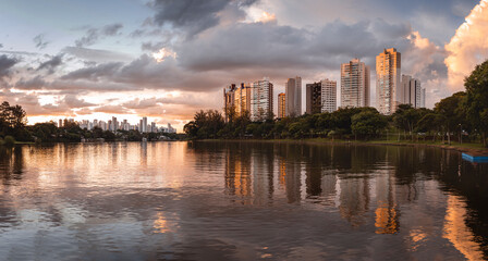 Fototapeta premium Urban scene at dusk, with tall buildings reflected in the city of Londrina, Brazil, surrounded by trees, under a partly cloudy sky.