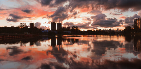 Naklejka premium Dramatic orange sky reflected in the calm waters of the lake in the city of Londrina, Brazil, while the silhouettes of buildings and trees create an urban setting at dusk.