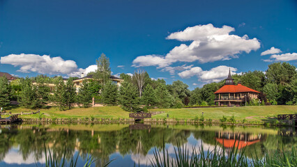 Fototapeta premium View of the pond, blooming meadow, forest and cottages in village at summer timelapse hyperlapse