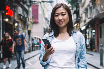 Cheerful Asian woman standing on street with smartphone and drink