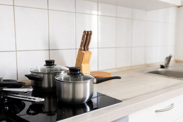 modern kitchen with an induction hob on which there are two metal pans with transparent lids. In the background there is a stand with knives and a neat tabletop, creating a feeling of order and cleanl