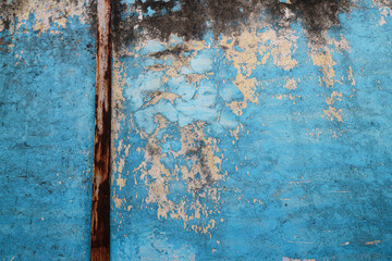 A close-up of a weathered blue wall with peeling paint and a rusty pipe. The texture is rough and aged, creating a vintage and rustic background. Cracked Rustic Blue Wall Texture Background.