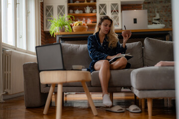 Obraz premium Young caucasian woman in spotted bathrobe enjoying morning read and breakfast in living room.