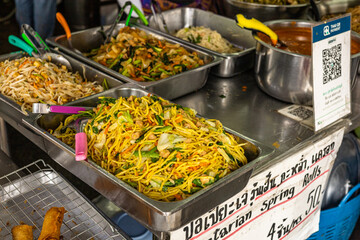 Street food scene in Thailand with a variety of delicious dishes like stir-fried noodles and spring rolls. A sign in Thai advertising 