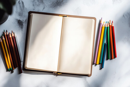 A sketchbook with blank pages on a light surface, with a set of colored pencils beside it, daylight, minimalistic composition, mockup with blank space.