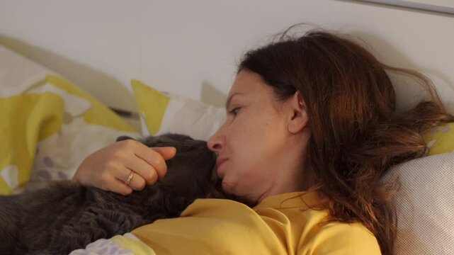 Happy relaxed woman lying in bed with cat at home, woman stroking her beloved domestic gray cat