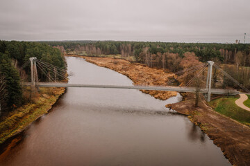 Aerial view of a suspension bridge crossing a calm river with surrounding autumnal trees, fields,...