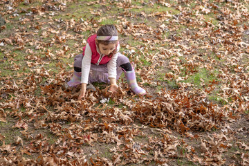 A young girl wearing a pink vest and rainbow boots collects autumn leaves in a park on a sunny afternoon, enjoying the seasonal beauty and engaging in playful exploration of nature.