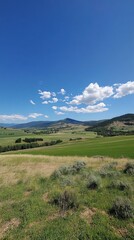 A scenic view of a lush green valley with mountains in the distance and a clear blue sky with white clouds.