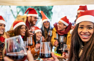 Smiling young people on santa hat celebrating Christmas with red wine at food street market - Winter friendship concept with friends enjoying time and having fun eating together at restaurant eve day