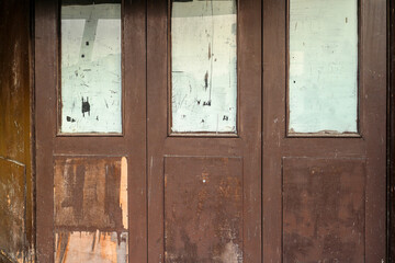 Close up of a weathered wooden door with faded glass panels. The door is made of brown wood, and the glass panels are covered in faded scratches. Old Wooden Doors with Faded Glass Panels.