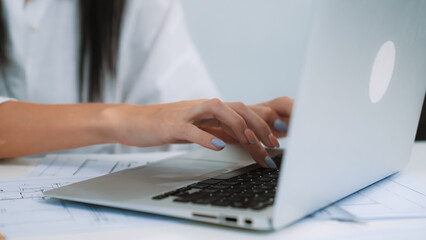 Close-up of young beautiful professional architect hands using laptop with blueprint and architectural document placed on table at modern office. Creative design concept. Focus on hand. Immaculate.