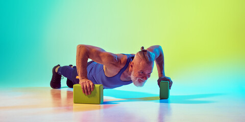 Athletic senior man in sportswear training, doing push ups yoga blocks against gradient green yellow background in neon light. Concept of sport, active elderly lifestyle, health care
