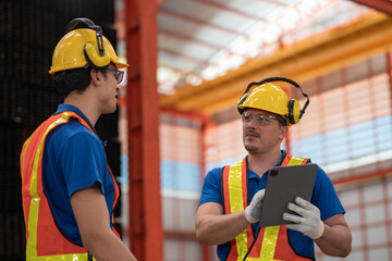 Two construction workers in safety gear, including yellow helmets and reflective vests, examine materials at industrial site. One holds a tablet, emphasizing digital tools in construction management.