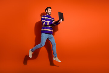 Full length photo of handsome young guy running hold netbook dressed stylish striped purple garment isolated on orange color background