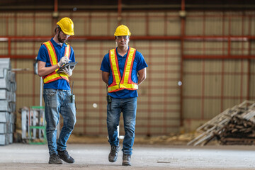 Two construction workers in reflective safety vests and helmets engage in conversation at industrial site. Both wear ear protection, emphasizing a noisy environment, with metal beams in the background