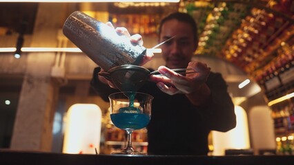 Closeup hand bartender using strainer mixed vodka in blue hawaii cocktail at luxurious counter. Preparation making special refresh drinks nightclub theme at nightclub on bokeh lighting warm. Vinosity.