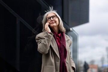 Fototapeta premium Elderly businesswoman talking on the phone outside her office building during a cloudy day