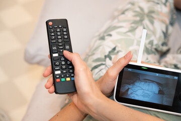 Mother relaxing on sofa holding tv remote and baby monitor capturing the balance between short moments of leisure and attentive caregiving, illustrating the life of a modern parent