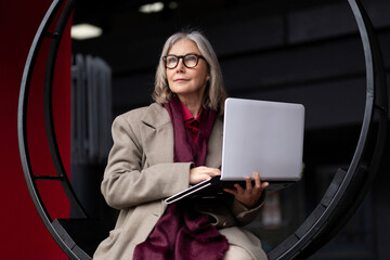 Elderly businesswoman working on a laptop in a modern urban environment during the afternoon