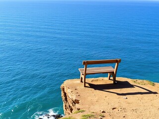 Fototapeta premium A weathered wooden bench sits alone on a cliff edge overlooking the vast expanse of the Pacific Ocean, with waves gently lapping at the base of the cliff below, natural beauty, serene landscape