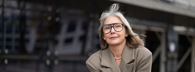 A senior businesswoman stands confidently outside a modern office building on a sunny day,...