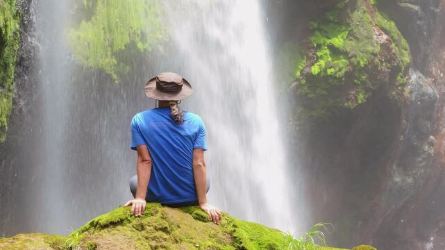 A young boy relaxes on mosscovered rocks, enchanted by a stunning waterfall in Costa Ricas lush landscape, enjoying a perfect escape into natures soothing tranquility