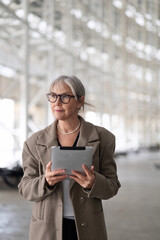 Stylish elderly woman in a chic outfit holds a tablet while exploring a spacious modern building interior during the daytime