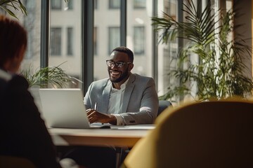 Smiling Man Working on a Laptop in a Modern Office With Large Windows and Greenery in the Early Afternoon