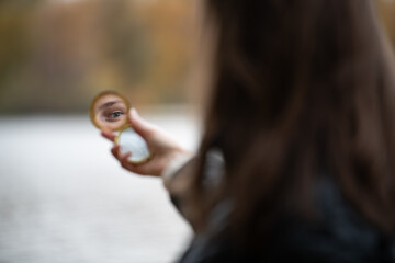 The subject gazes into a small mirror, capturing their eye, with a tranquil lake and vibrant autumn leaves in the background, creating a reflective moment. Focus on the reflection in the mirror.