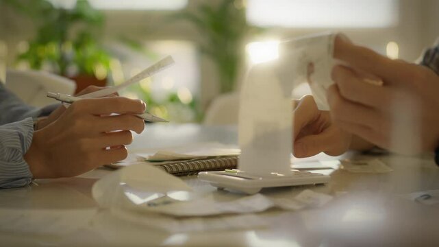 Individuals sort through numerous receipts while using a calculator to manage their finances in a sunny indoor setting. The atmosphere feels collaborative and focused on budgeting