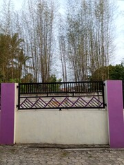 A modern fence with a purple accent wall and pillars. The fence features a geometric design and is situated in front of a bamboo grove.
