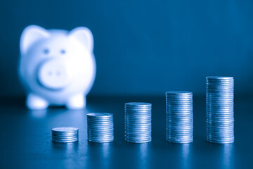 Stacks of Coins Representing Wealth and Financial Growth and white piggy bank in the background with blue color filter. Saving money and financial concept. 