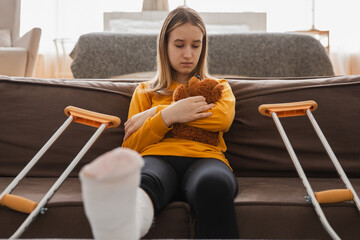 Young girl with broken leg cast sitting on sofa. Sad little kid hugging her teddy bear recovering...