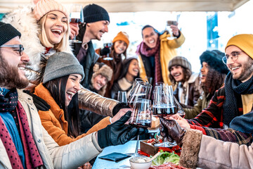 Group of young people toasting red wine at restaurant patio - Happy multiracial friends wearing winter clothes having fun at winery bar table - Dining life style and friendship concept