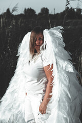 Brunette with big white angel wings and white casual clothes stands in nature against the backdrop of tall grass and trees and a clear blue sky. Angel girl descended to earth.