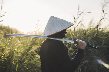 A samurai in traditional clothing carries a katana at sunset in a field bathed in golden light,...