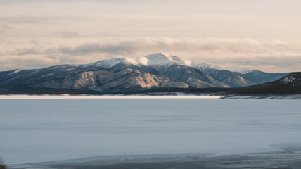 Obraz premium A serene winter landscape featuring a majestic mountain with snow-capped peaks, reflecting softly on the frozen lake under a golden sky.