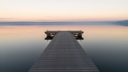 Obraz premium A serene wooden dock extends into calm waters at dusk, creating a tranquil scene with soft pastel colors reflecting on the surface.